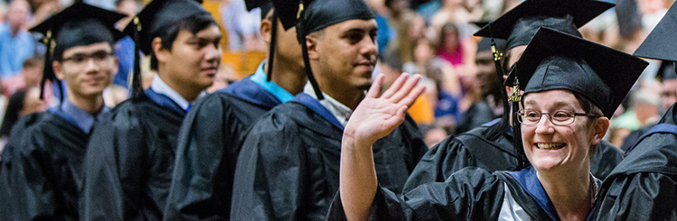 Group of graduates standing in line to get diploma, one graduate waving to friend/family member