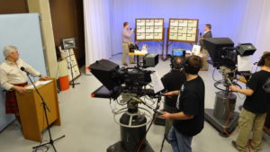 Media Arts and Technology program volunteers working the camera capturing the Rotary auction host behind a podium and two other hosts presenting the items