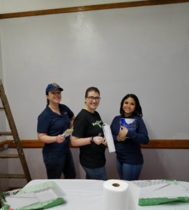 Three people stand in front of a white wall holding painting supplies.