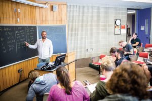 A professor stands in front of a classroom of students as he points at a whiteboard covered with financial figures.