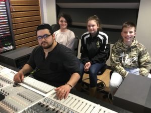 College student sitting in front of the mixing board with three middle school students behind him