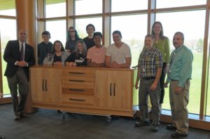 A group of students and school administrators stand in front of a piece of furniture that is roughly 3 feet tall, 12 feet long and with multiple drawers.