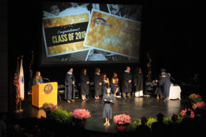 A student holds her diploma on stage with other people standing at the back of the stage.