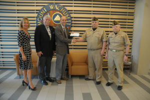 Five people stand in a row with the middle two holding a check and shaking hands.