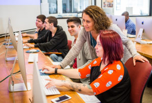 A class of students in a Mac computer lab, focus on instructor and student looking at a computer