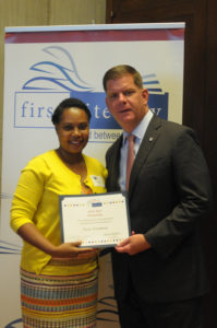 A woman in yellow stands next to Boston's mayor as they both hold up a certificate.