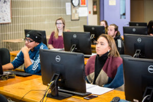 Students sit at computers in a classroom space as they face forward.