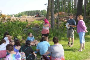 A man stands in front of a group of students speaking to them. They are outside standing in front of an area of cleared woods.