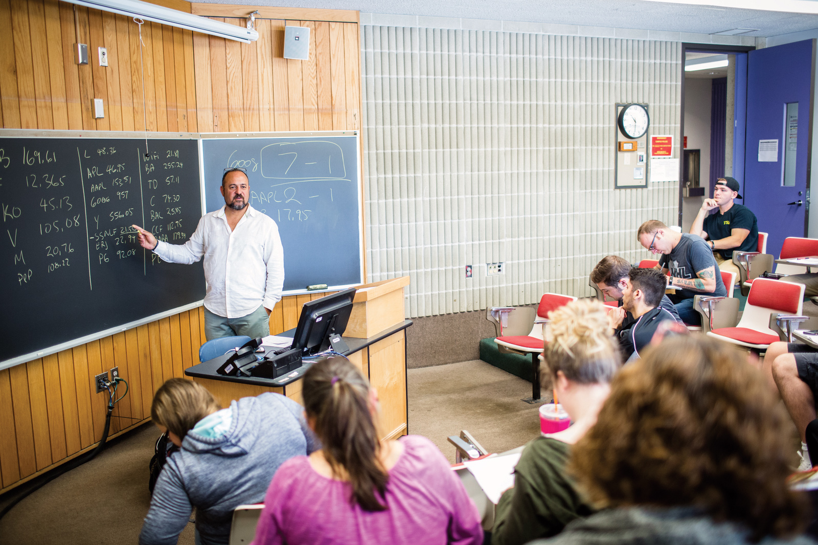 A male professor stands at the front of a classroom in front of a chalkboard facing students seated in stadium-style seats climbing away from him.