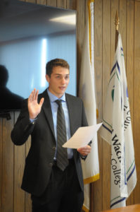 A man in a suit holds his right hand up while holding a paper with his left hand.