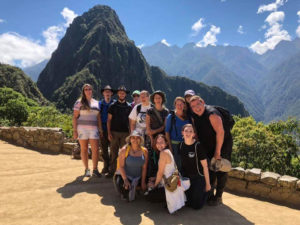 A group of people poses on a stone pathway with soaring mountains in the background.