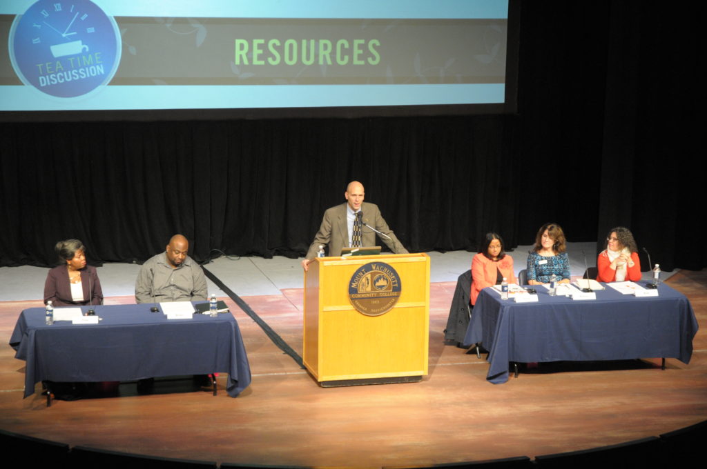 A man stands at a podium on a stage flanked by two tables with speakers sitting at those tables.