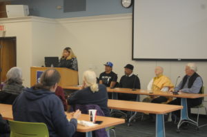 Four people sit at a table in the front of a room giving a presentation. To the left is the moderator at a podium.