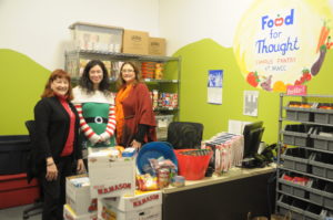 Three people stand in the MWCC food pantry in front of shelves full of food and a desk piled with food.