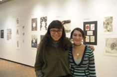 Two students stand next to each other in front of a white walled gallery space with artwork hanging on the walls.