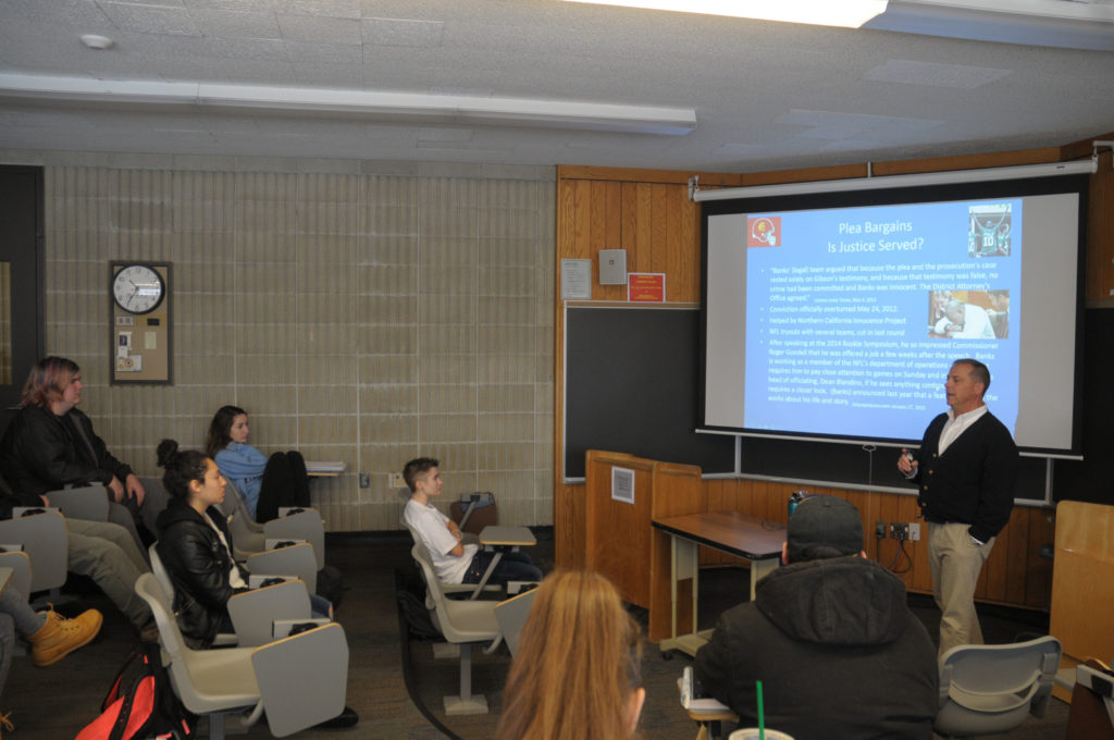 A person stands in front of a screen teaching a classroom of students who are seated.