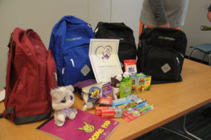 Three backpacks are lined up with school supplies and snacks in front of them.