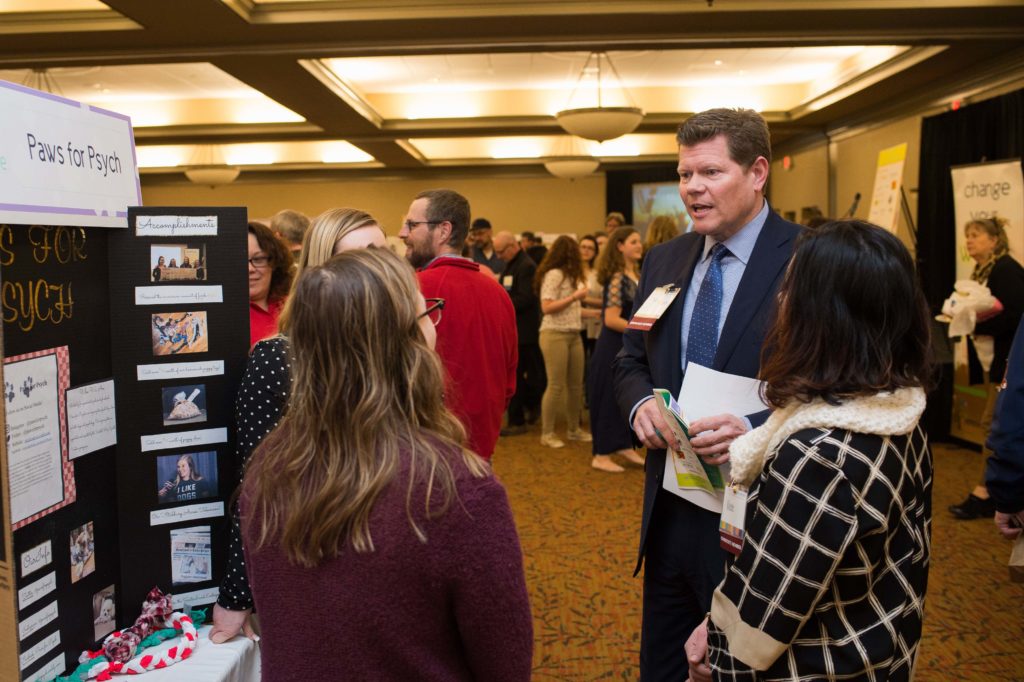 A person talks to two students in front of a poster board display. A crowd of people is shown milling around in the background.