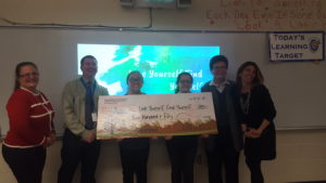 2 kids and 4 adults stand in front of a slideshow while holding a giant check