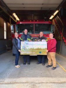 1 teen and 1 adult stand behind 2 firefighters and a firetruck while holding a giant check