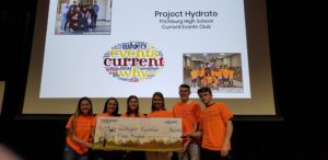 6 teens stand in front of a slideshow while holding a giant check