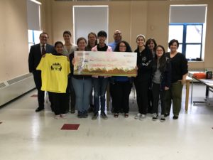 6 teens and 6 adults stand in line behing a giant check, one is holding a t-shirt