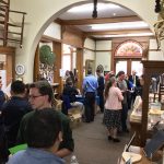 A group of around 20 people mingles in an entry room at an older house with historical artifacts hanging on the walls and in a case.