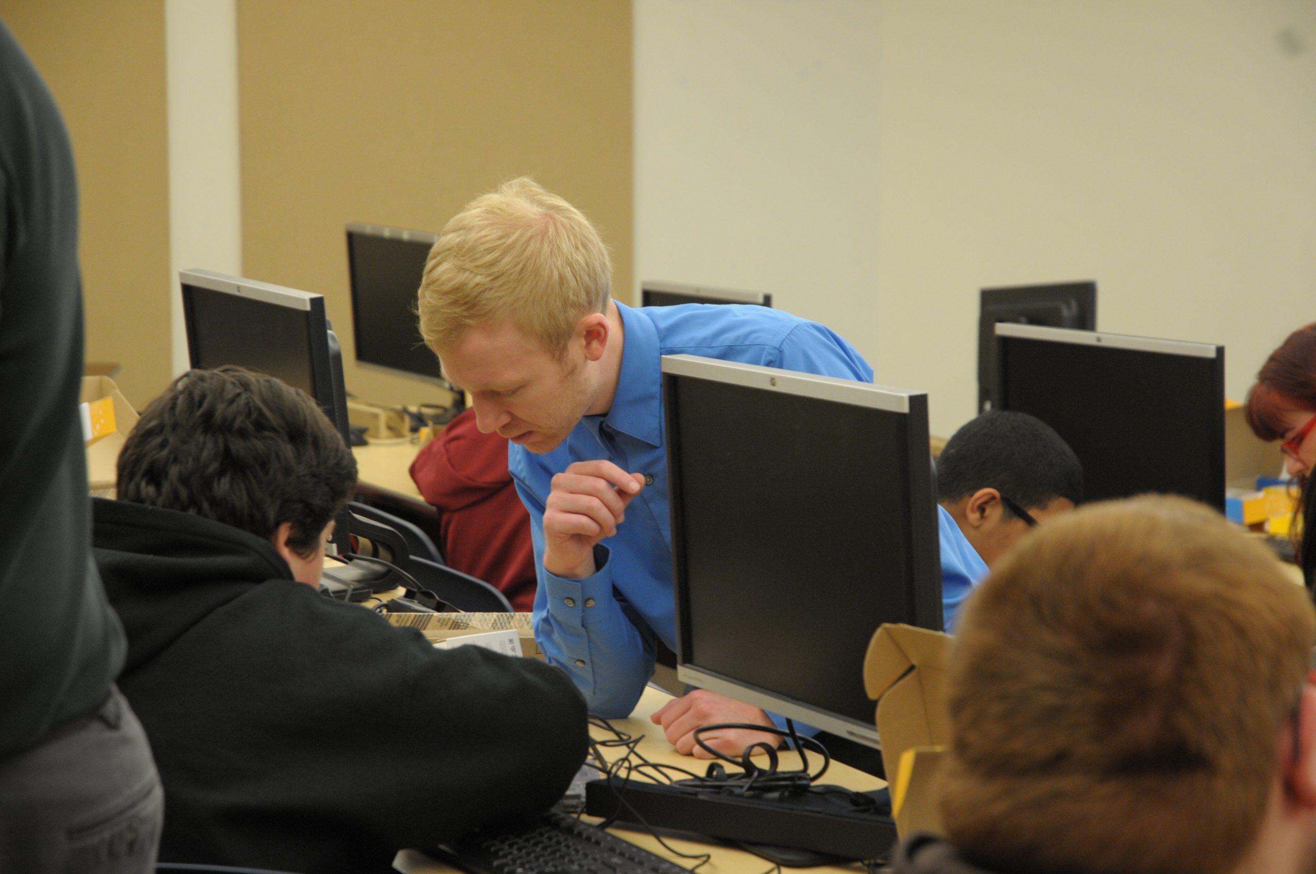 People work in front of computers as one person leans over helping one individual.