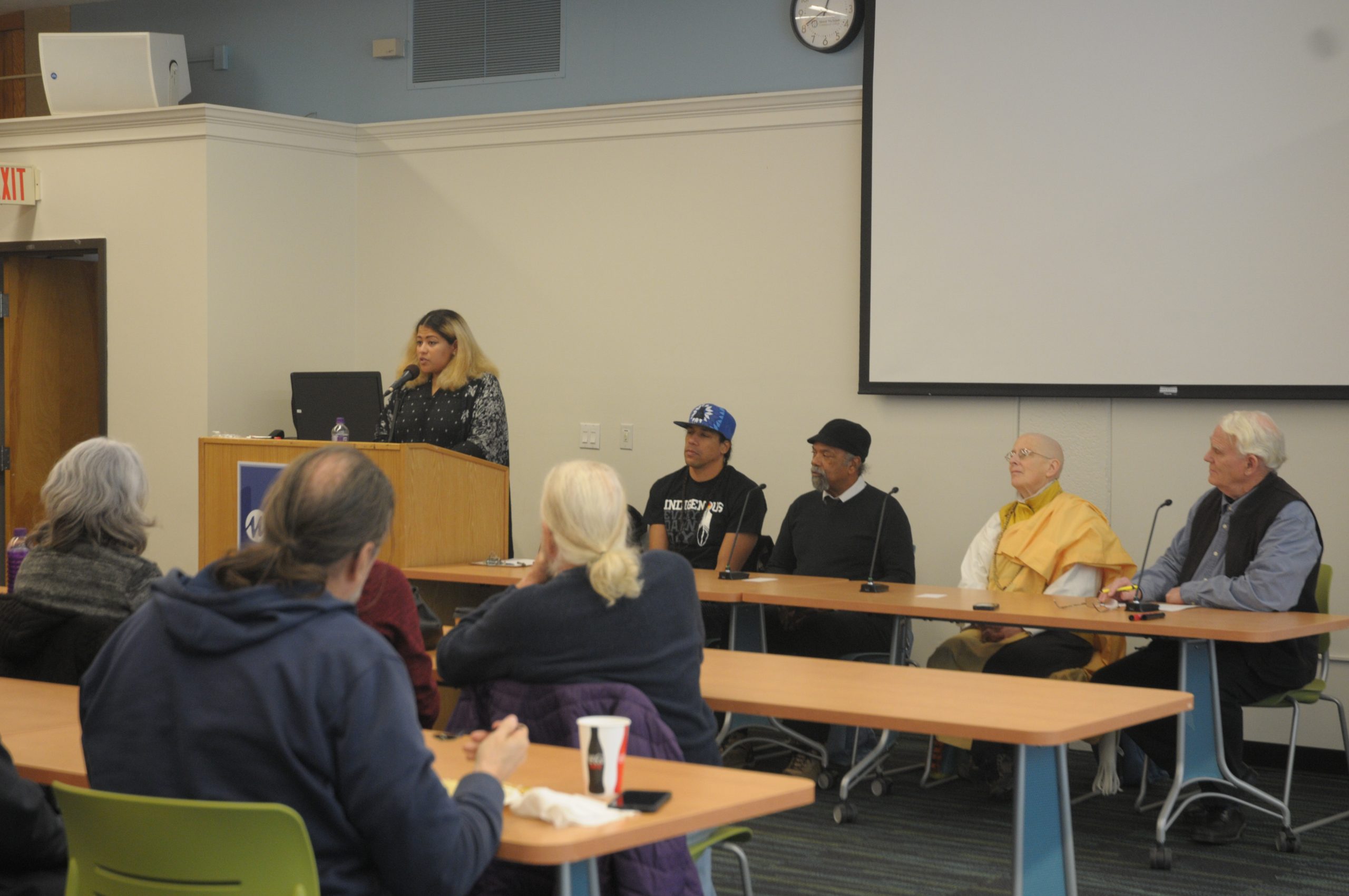 Four people sit at a table in the front of a room giving a presentation. To the left is the moderator at a podium.