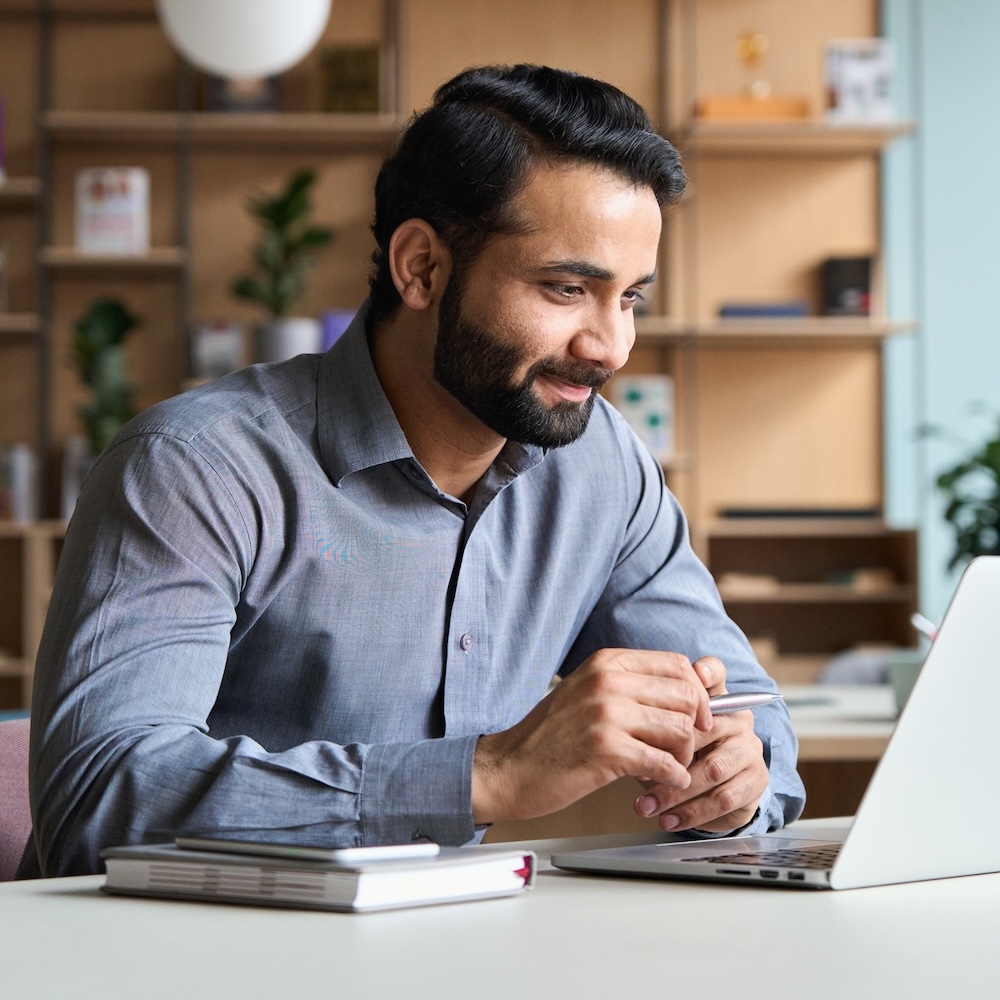 Smiling bearded indian businessman working on laptop at home office. Young indian student using computer remote studying, virtual training on video call meeting, watching online webinar or seminar.