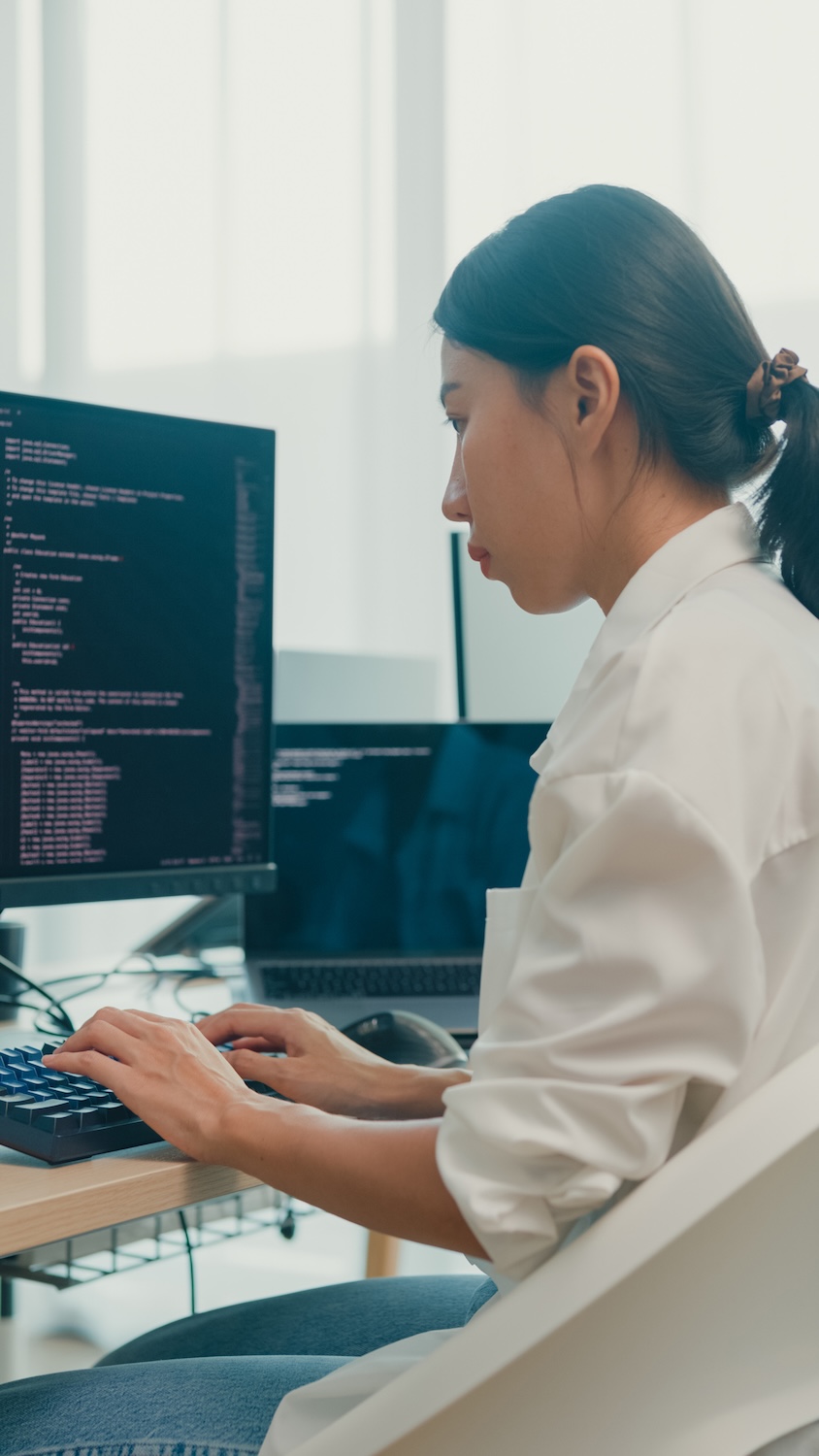 Young Asian woman software developers using computer to write code sitting at desk with multiple screens work at office. Programmer development. Vertical Screen.