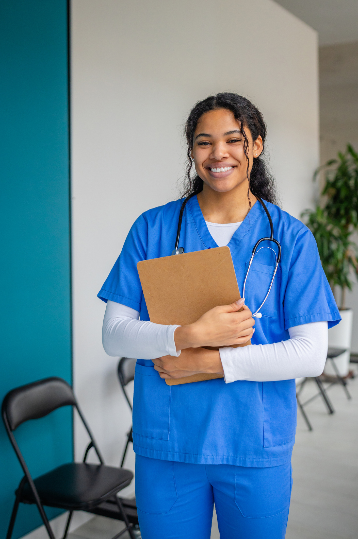 Healthcare professional smiling in a hospital waiting room while holding a clipboard and wearing medical scrubs