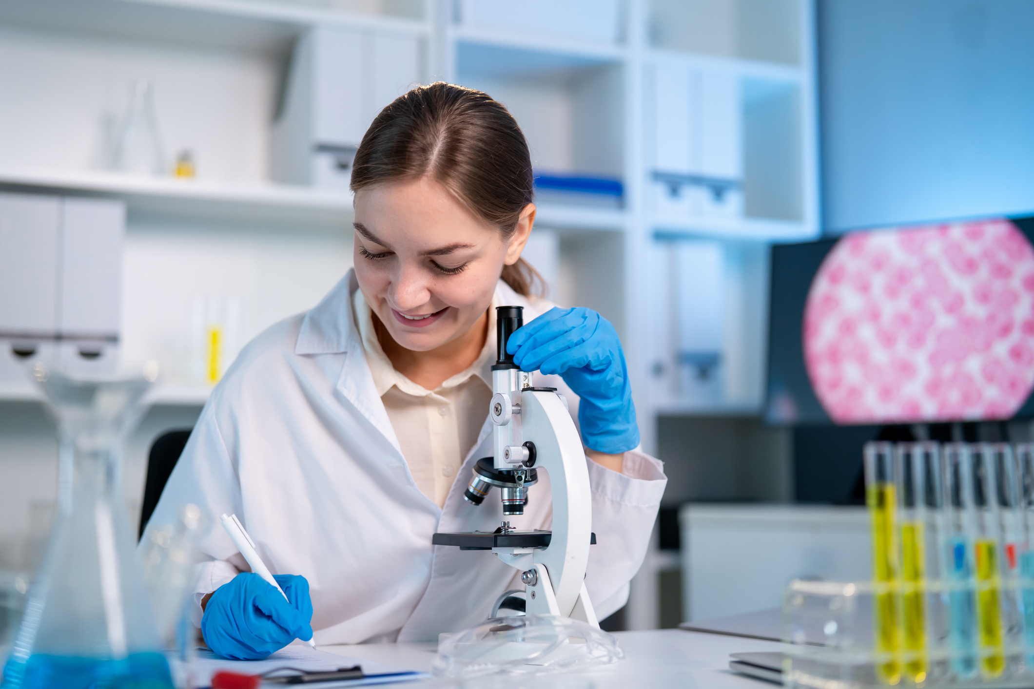 Caucasian female scientist in white lab coat working in the laboratory. Attractive young researcher technician sitting at a desk, taking notes and conducting medical research in modern science lab.