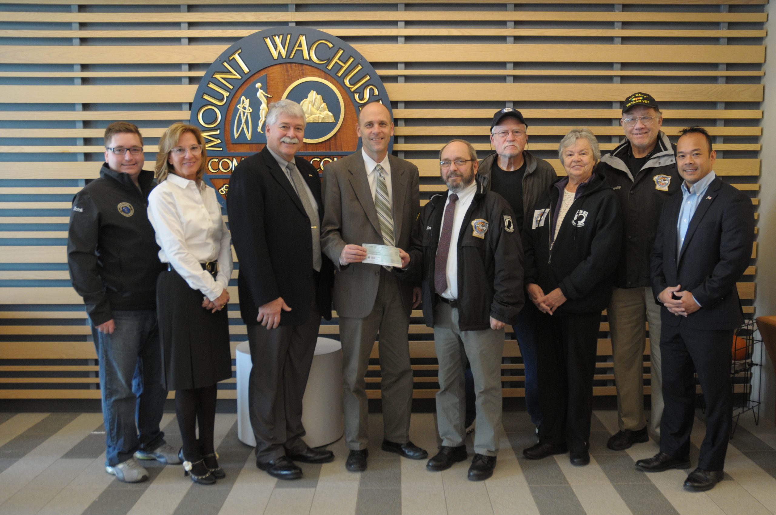 People stand in a line in front of the college's seal during a check donation ceremony.