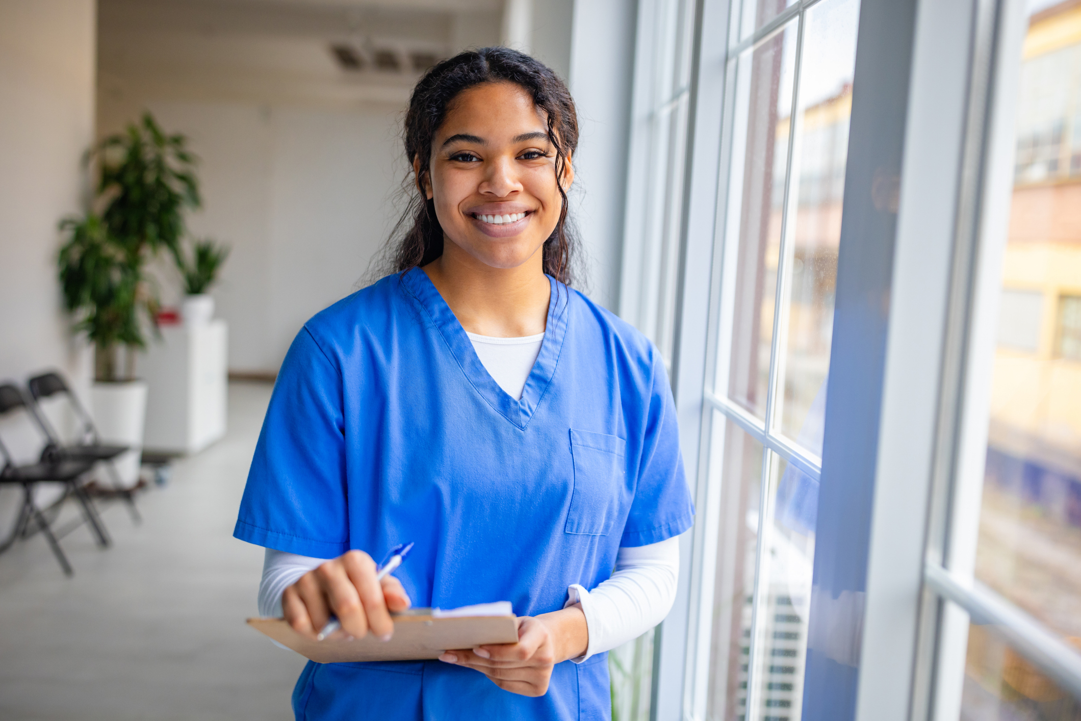 Smiling female nurse in scrubs takes notes while standing in a hospital waiting room during a busy shift