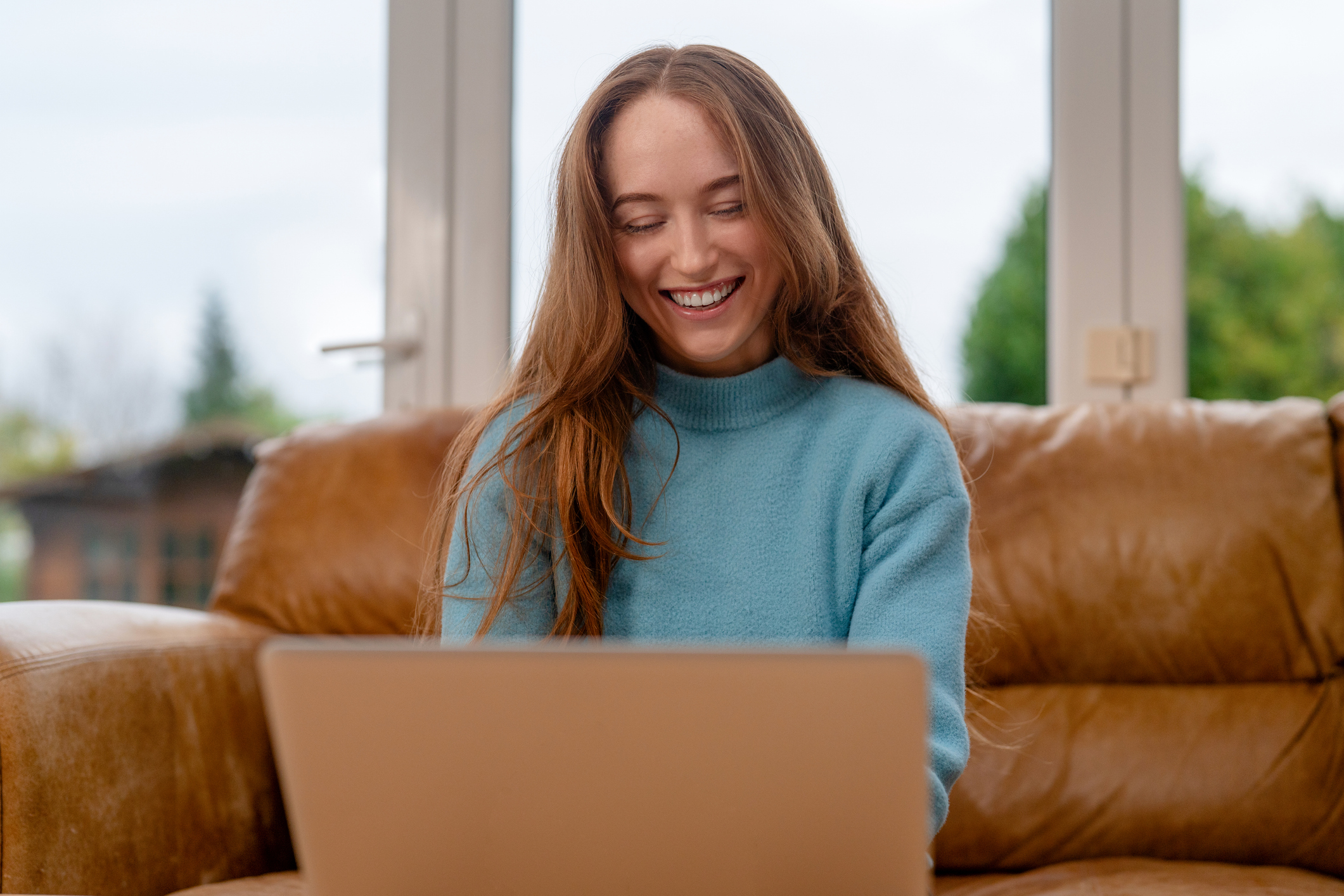 Woman sits on couch and smiles while using laptop at home in the afternoon