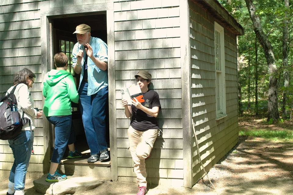 Three faculty walking into Walden's cabin while a student reads the book outside next to the door