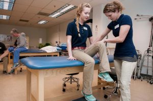 One female student sitting on a table while another female student checks the range of motion in her bent knee